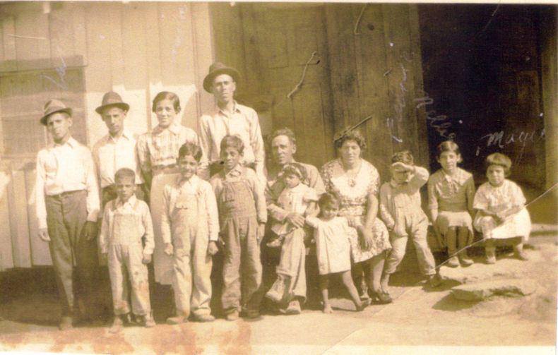 Back Row: standing against the house, left to right, Jack, Jim, Juanita and Joe Gomez; front row: 
 left to right Sammy, David, Gene, father George Gomez holding Mary in his lap, Rosie bare foot, mother Susana Gomez, Freddy, Nelly and Maggie.  Susie Gomez
